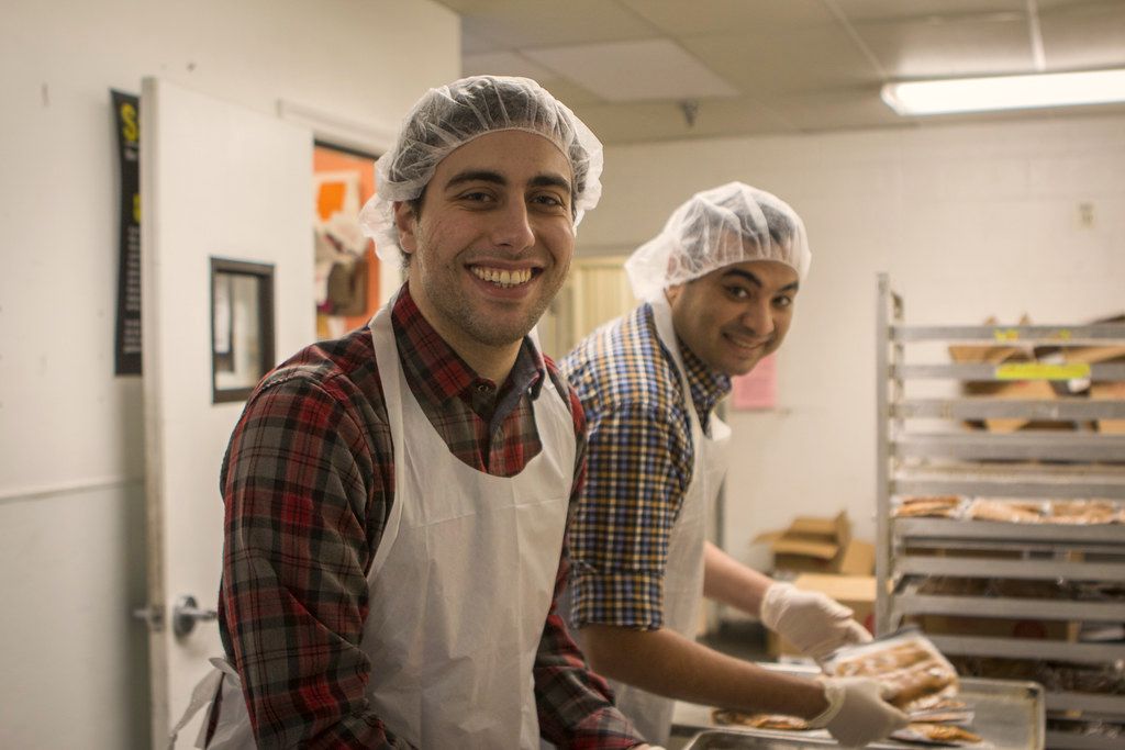 Two smiling volunteers in aprons and hair nets carrying trays of food past tall kitchen racks
