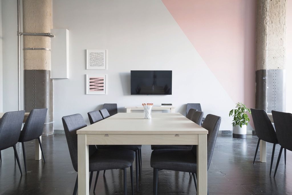 A calm, empty meeting room with dark chairs, a pale wooden table and one soft-pink wall