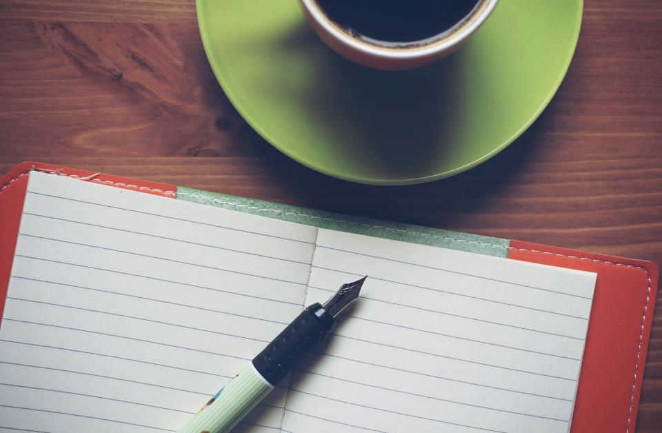 An open notebook with a fountain pen resting on it, beside a black coffee on a citron-green saucer