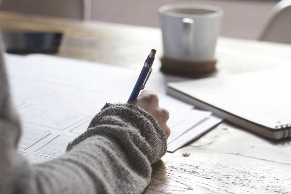 A hand writing notes at a wooden desk beside a mug of coffee, in soft window light