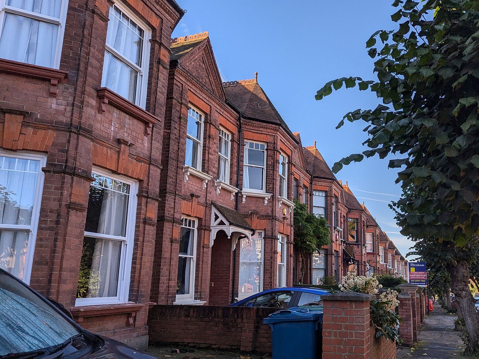 A tree-lined row of red-brick Victorian terraced houses with bay windows and white sash frames in low evening sun