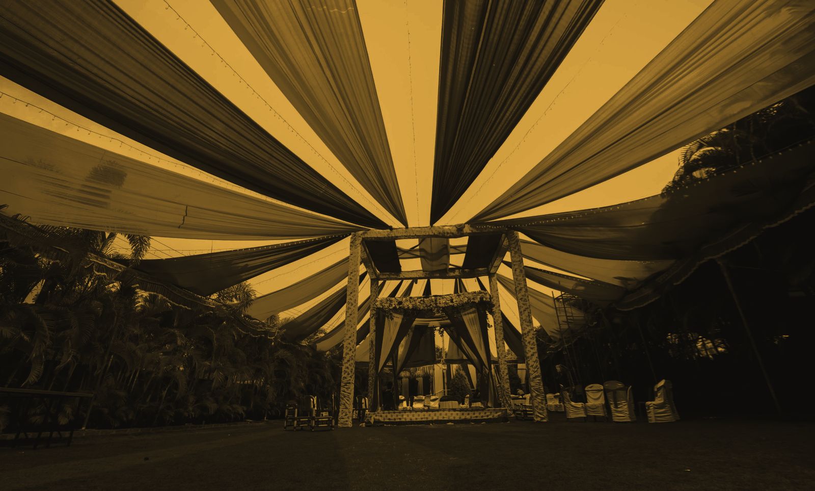 An outdoor mandap at dusk, canopy drapes radiating overhead, chairs still empty
