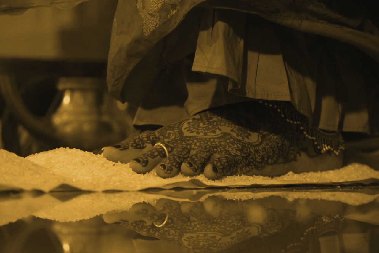 A bride’s mehndi-covered feet stepping onto rice during the sapthapadi, reflected in the polished floor