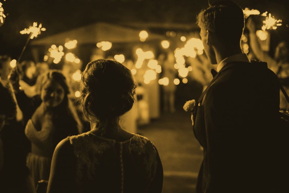 A couple seen from behind, walking into a crowd of guests holding sparklers at night