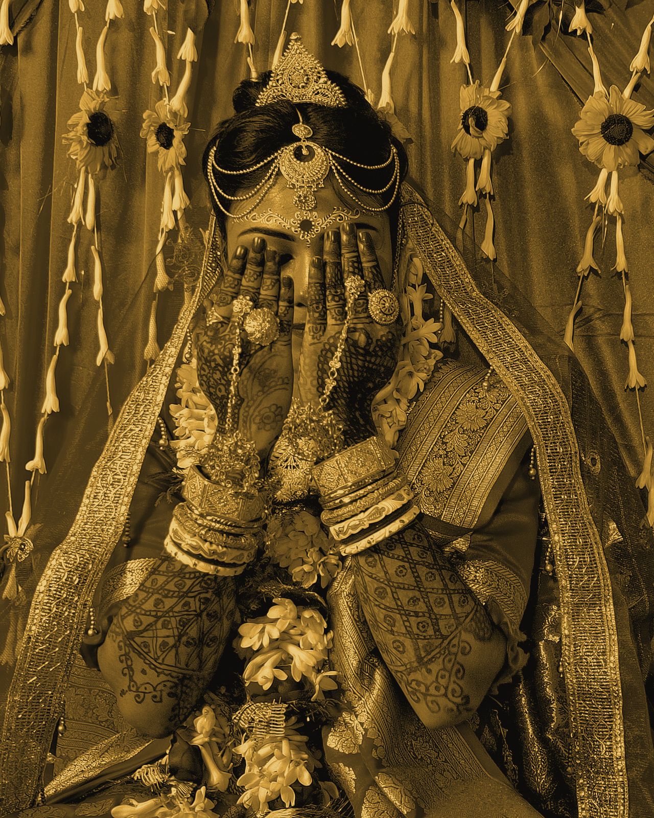 A bride covering her face with henna-covered hands, framed by hanging flower garlands