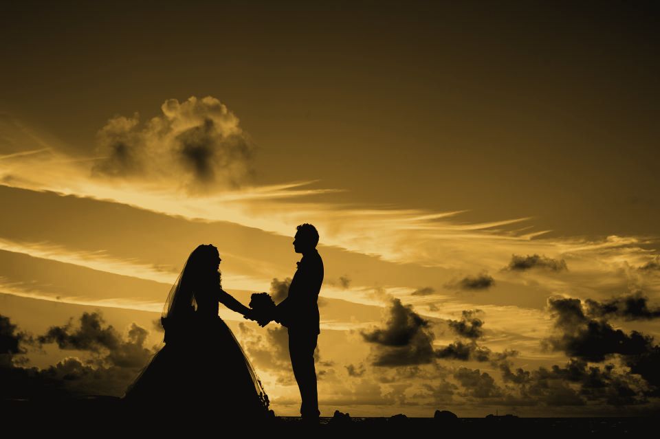 A couple in wedding clothes holding hands in silhouette against a sunset sky