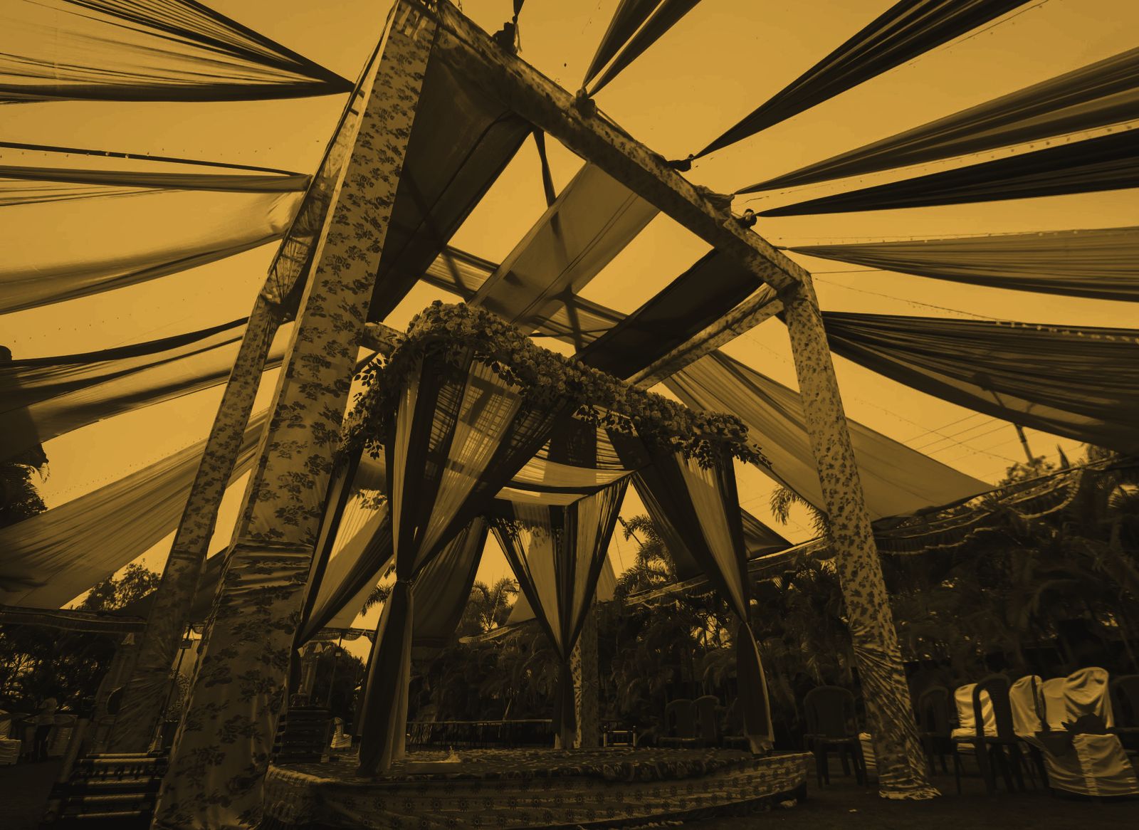A pillared mandap under radiating canopy drapes at dusk, garlanded and empty before the guests arrive